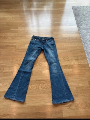 True religion jeans - Säljer ett par blå jeans med låg midja och rejält utsvängda ben. Klassisk femficksmodell med snygga kontrastsömmar och slitningar framtill. Perfekta för dig som gillar retrostil och vill sticka ut med en cool siluett. Använda 1 gång