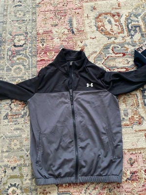 Svart och grå träningsjacka Under Armour - Snygg träningsjacka från Under Armour i svart och grått. Jackan har hel dragkedja, hög krage och två sidofickor. Tillverkad i ett lätt och stretchigt material som passar perfekt till träning eller uppvärmning. Under Armour-logga på bröstet.