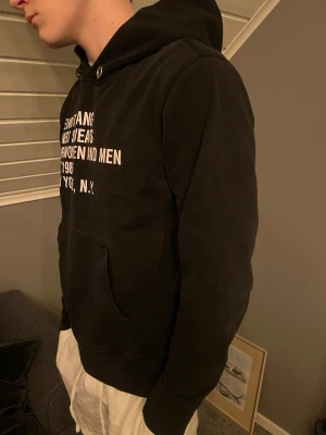 Svart hoodie från Helmut Lang - Säljer en svart hoodie från Helmut Lang med vitt tryck på bröstet. Hoodien har luva och en  magficka. Materialet är mjukt och skönt, perfekt för en avslappnad stil. Texten på framsidan ger en cool och modern vibe. Storlek L men sitter som en M. 
