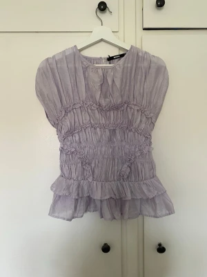 Ljuslila volangblus med rynk - Supersöt ljuslila blus med volanger och rynkade detaljer. Toppen har korta ärmar, rund halsringning och är gjord i ett tunt, skirt material som ger en luftig känsla. Perfekt för dig som gillar romantisk och trendig stil. St L men passar M. 