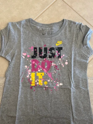 Grå Nike t-shirt med färgstänk - Snygg grå t-shirt från Nike med trycket 'JUST DO IT.' i svart, rosa och gult samt färgstänk i rosa och vitt. Klassisk rund halsringning och korta ärmar. Perfekt för en sportig och avslappnad stil.
