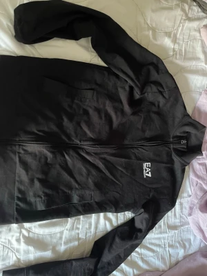 Svart zip hoodie från EA7 - Säljer en svart hoodie med dragkedja från EA7 Emporio Armani. Hoodien har två fickor framtill och EA7-logga på bröstet samt ett vitt tryck på ryggen. Tillverkad i mjukt material och har en klassisk passform med långa ärmar.