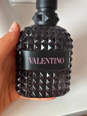 Valentino parfym med nitar - Säljer en lyxig parfym från Valentino i en mörk, nitmönstrad glasflaska med svart och rosa detaljer. Flaskan har en unik, edgy design med pyramidformade nitar och svart kork. Perfekt för dig som gillar exklusiva och snygga parfymer.  Är fylld upp till där mina fingrar slutar. Fullständiga namn är Valention Uomo Born in Roma Edt. Köptes för 1080 kr