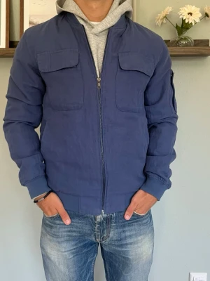 Dressmann Linneblandningsjacka – Marinblå | Bomber | Strl S - DRESSMANN LINEN BLEND JACKA  Storlek: S Passform: Regular bomber-fit Färg: Marinblå Material: Linen blend (linneblandning)  Lätt och luftig jacka i linneblandning — perfekt för vår och tidig höst. Dubbla bröstfickor och armficka ger den ett sportigare, mer premium-inspirerat uttryck. Dragkedja i marinblå som matchar tyget snyggt.  Modellen på bilden är 175 cm och väger 65 kg — jackan sitter som en S ska sitta.  Skick: Mycket bra skick utan defekter.