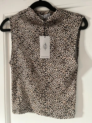Ärmlös leopardmönstrad topp - Snygg ärmlös topp från Stradivarius med leopardmönster i beige, svart och brunt. Toppen har hög hals och är tillverkad i ett stretchigt, tunt material som sitter tajt mot kroppen. Perfekt för att skapa en trendig och cool look.