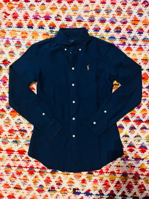 Mörkblå skjorta från Ralph Lauren XS - Klassisk mörkblå skjorta från Ralph Lauren i bomull. Skjortan har lång ärm, vita knappar framtill och broderad logga på bröstet. Snygg passform med button-down krage och tidlös design som funkar till mycket.
