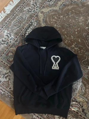 Ami Paris hoodie - Säljer en svart hoodie med huva och dragsnören. På bröstet finns en stor vit patch med ett hjärta och bokstaven A. Hoodien har en klassisk passform och är tillverkad i mjukt material, perfekt för en chill och stilren look.
