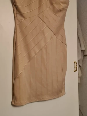 Beige bandageklänning med struktur - Snygg beige bandageklänning med figurnära passform och markerade sömmar i olika riktningar som ger en cool struktur. Klänningen är ärmlös och har rundad urringning både fram och bak. Materialet är stretchigt och tjockt för en smickrande siluett.