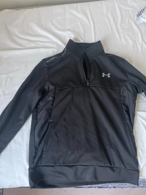 Svart träningströja från Under Armour - Snygg svart långärmad träningströja från Under Armour med halv dragkedja framtill och logga på bröstet. Tröjan har hög krage och diskreta fickor med dragkedja. Materialet är funktionsmaterial som passar perfekt till träning.