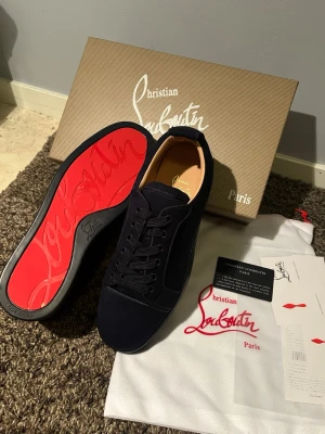 Svarta Christian Louboutin sneakers - Snygga svarta sneakers från Christian Louboutin med klassisk röd sula och ovandel i mocka. Skorna har svarta skosnören, diskret design och exklusiv känsla. Perfekta för dig som vill sticka ut med en lyxig detalj. Kommer med originalkartong och dustbag.