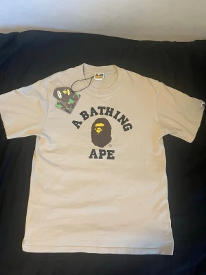 Beige A Bathing Ape t-shirt - Säljer en beige t-shirt från A Bathing Ape med det klassiska BAPE-logotrycket i svart och gult på bröstet. T-shirten har rund hals och korta ärmar. Perfekt för dig som gillar streetwear och vill sticka ut med en ikonisk design.