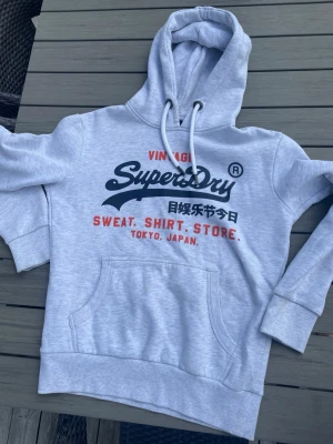 Ljusgrå Superdry hoodie med tryck - Ljusgrå hoodie från Superdry med stor logga och text i svart och rött på bröstet. Klassisk känguruficka framtill, justerbar huva med vita snören och ribbade muddar. Perfekt för en avslappnad streetwear-look. Kan går ner i pris vid snabb affär. Hör av dig vid frågor