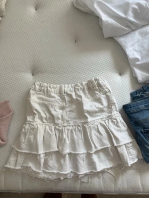 Vit volangkjol  - Supersöt vit kortkjol med flera volanger och råa kanter för en avslappnad look. Kjolen har fickor fram, bälteshällor och elastisk midja för extra komfort. Perfekt för dig som gillar en trendig och ungdomlig stil.