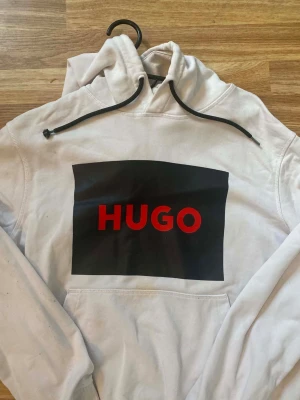 Hugo boss hoodie - Säljer en vit hoodie från HUGO med stor svart ruta och röd HUGO-text på bröstet. Hoodien har huva med svarta snören och en klassisk magficka. Perfekt för dig som gillar streetwear och vill ha en enkel men snygg look.