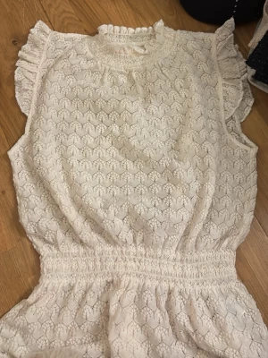 Vit spetsblus från Holly & Whyte - Söt vit spetsblus från Holly & Whyte by Lindex med volangdetaljer vid axlarna och halsen. Blusen har ett hjärtformat spetsmönster, resår i midjan och liten volangkrage. Perfekt för dig som gillar romantisk stil.
