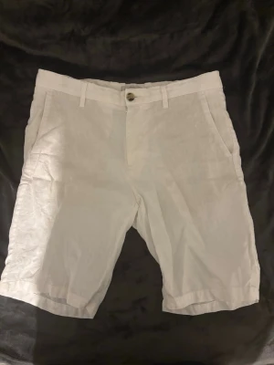 Vita linneshorts med fickor - Snygga vita shorts i linne med klassisk passform. De har bälteshällor, sidofickor fram och två bakfickor med knapp. Perfekta för varma dagar och ger en fräsch och stilren look.