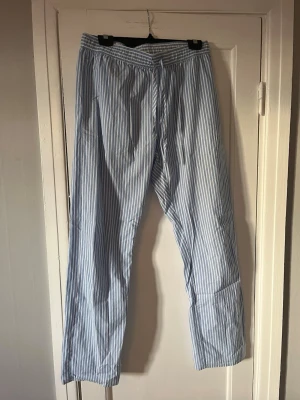 Blåvitrandiga pyjamasbyxor - Säljer ett par klassiska pyjamasbyxor med blå och vita vertikala ränder. Byxorna har resår och dragsko i midjan för skön passform. Tillverkade i ett mjukt och lätt tyg, perfekta för chill hemma eller sovdags.