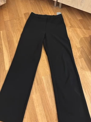 Svarta kostymbyxor från Vero Moda - Snygga svarta kostymbyxor från Vero Moda med raka, vida ben och klassisk midja. Byxorna har bälteshällor och är tillverkade i ett mjukt, vävt material som ger en stilren look. Perfekta för dig som gillar en clean och modern stil.