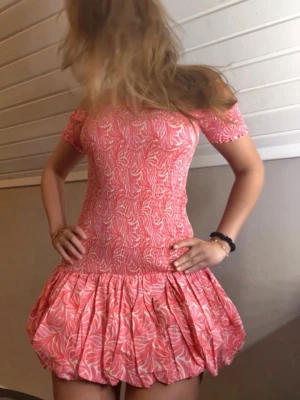 Rosa offshoulder kortklänning - Supersöt kortklänning i rosa och vitt med offshoulder och puffig kjol. Klänningen har ett mönster med vita blad, smockad överdel och volang nedtill. Perfekt för sommarens alla tillfällen och ger en riktigt trendig vibe.