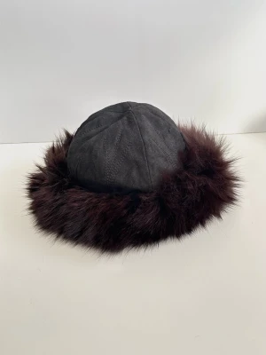 Cronquists - Svart hatt / mössa med brun päls - storlek M / 57 - Härlig hatt / mössa i 100% äkta mocka och päls från anrika körsnären / pälstillverkaren Cronquist i storlek M / 57 (inre omkrets 57 cm). Tidlös modell med formsydd rundad topp i mocka / läder och med en mjuk och fluffig kant i päls. Hatten har en rundad form med dekorativa sömmar på ovansidan. Insidan är fodrad med ett quiltat tyg för extra komfort. Fint skick!