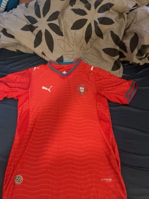 Röd Portugal fotbollströja  - Snygg röd Portugal fotbollströja med landslagets emblem på bröstet. Tröjan har gröna detaljer vid krage och ärmslut samt diskret vågmönster i tyget. Perfekt för match eller träning.