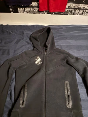 Svart AIK hoodie med dragkedja - Säljer en svart hoodie med AIK-logga på bröstet och stor AIK-text på ryggen. Hoodien har dragkedja framtill, två fickor med dragkedja och en stor huva. Perfekt för dig som vill visa ditt AIK-support. Materialet känns mjukt och bekvämt.