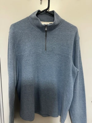 Blå stickad tröja med halv dragkedja - Mysig blå stickad tröja från RW&CO. med halv dragkedja framtill och hög krage. Tröjan har lång ärm och är tillverkad i ett mjukt material som passar perfekt för kyligare dagar. Enkel och stilren design som är lätt att matcha.