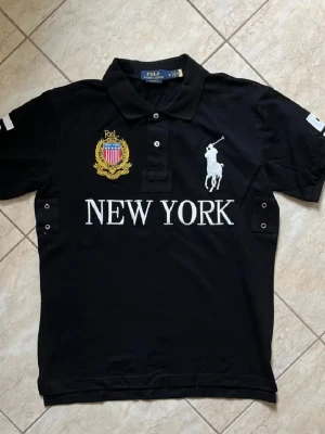 Svart Polo Ralph Lauren new york - Svart pikétröja från Polo Ralph Lauren med stor vit logga, broderad New York-text och emblem på bröstet. Nummer 5 i vitt på ryggen och detaljer på ärmarna. Klassisk krage och knappar framtill. Perfekt för en sportig och stilren look.