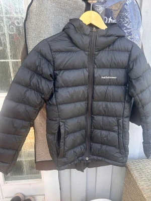 Svart dunjacka Peak Performance - Snygg svart dunjacka från Peak Performance med huva och dragkedja framtill. Jackan har en puffig design med horisontella sömmar och logga på bröstet. Perfekt för kalla vinterdagar och riktigt skön att bära. I bra men använt skick med 2 lagningar, se bild.