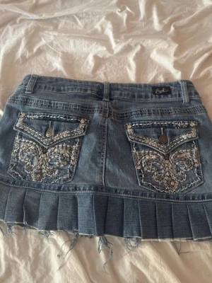 Blå jeanskjol med broderade fickor - Säljer en blå jeanskjol med plisserad nederkant och slitna detaljer. Kjolen har två stora bakfickor med snygga broderier och knappar, samt klassiska framfickor. Perfekt för dig som gillar en cool och avslappnad stil.