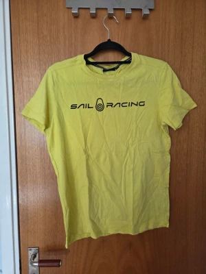Gul Sail Racing t-shirt - Gul t-shirt från Sail Racing med svart logga och text på bröstet samt en liten logga i nacken. Klassisk passform och rund hals. Perfekt för dig som gillar enkel och sportig stil.