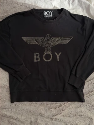 Svart sweatshirt från BOY London - Svart sweatshirt från BOY London med ikonisk örnlogga och texten BOY framtill. Tröjan har rund halsringning, långa ärmar och muddar. Perfekt för en streetwear-look och passar till det mesta i garderoben.
