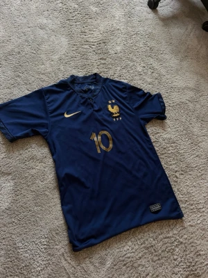 Frankrike T shirt storlek 176 - Mörkblå fotbollströja med guldiga detaljer, nummer 10 och Mbappé på ryggen. Tröjan har korta ärmar, två stjärnor ovanför Frankrikes landslagsemblem och Nike-logga på bröstet. Perfekt för dig som älskar fotboll och vill stötta Frankrike.