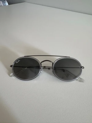 Solglasögon från Ray-Ban - Snygga runda solglasögon från Ray-Ban med tunn metallbåge i silver och mörka glas. Klassisk design med dubbla bryggor och diskret Ray-Ban-logga på linsen. Perfekta för dig som vill ha en tidlös och trendig look.