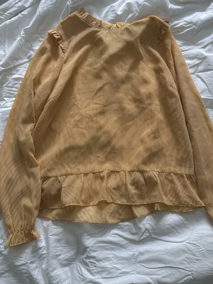 Gulrandig blus med volangdetaljer - Söt blus i ljusgul och vit med tunna ränder och volangdetaljer vid hals, ärmslut och nederkant. Blusen har långa ärmar och en liten knappöppning bak i nacken. Tillverkad i ett lätt och luftigt tyg, perfekt för en trendig vardagslook.
