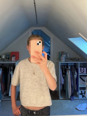 Ljusgrå stickad kortärmad tröja - En enkel och stilren ljusbeige stickad tröja med korta ärmar och rak passform. Tröjan är mjuk och har en något croppad längd, perfekt till jeans eller kjol. Materialet ser ut att vara en ull- eller bomullsblandning.