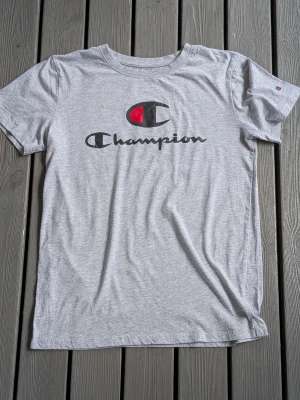 Grå Champion t-shirt med logga storlek M - Snygg grå t-shirt från Champion med klassisk logga på ärmen och stort tryck framtill. Tillverkad i mjuk bomulls- och polyestermix som känns skön mot huden. Perfekt till en avslappnad streetwear-look. Storlek M.