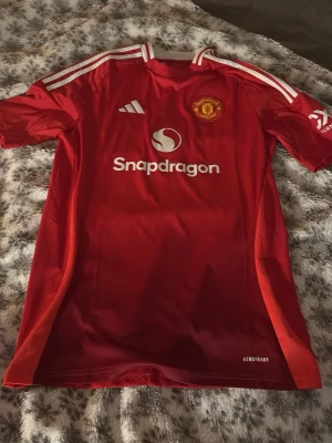 Manchester United matchtröja röd Adidas - Säljer en röd Manchester United matchtröja från Adidas med vita detaljer och klubbmärke på bröstet. Tröjan har Snapdragons logga på framsidan och klassiska Adidas-ränder på axlarna. Tillverkad i lätt och ventilerande material, perfekt för fotbollsträning.