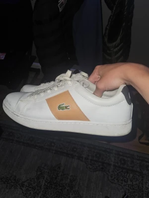Vita Lacoste sneakers med beige detalj - Snygga vita sneakers från Lacoste med beige panel på sidan och klassiska gröna krokodilloggan. Skorna har vita snören, svart detalj med vit Lacoste-logga på hälen och är tillverkade i slätt läder. Perfekta för en clean och stilren look.