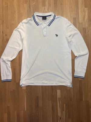 Vit långärmad Paul Smith piké med blå detaljer - Vit långärmad pikétröja med blå ränder på krage och ärmslut. Klassisk krage med knappar och en liten broderad zebra på bröstet. Perfekt för en clean och stilren look.