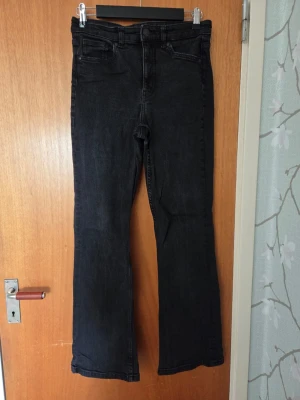 Svarta bootcut jeans med hög midja - Snygga svarta jeans med bootcut passform och hög midja. Jeansen har klassiska fem fickor och dragkedja med knapp framtill. Perfekta för dig som gillar en retroinspirerad look och vill ha ett par jeans som sticker ut lite extra.