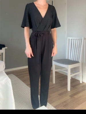 Svart jumpsuit med omlottopp - Snygg svart jumpsuit med omlottopp och vida korta ärmar. Byxorna är raka med hög midja och knytskärp. Toppen har lätt glittrig finish som ger en extra touch. Perfekt för dig som gillar stilrena och trendiga plagg. Använd en gång så är i nyskick. Passar även stl 36