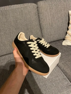 Maison Margiela skor - Stilrena svarta sneakers med beige sula och vita snören. Ovandelen är i en mix av mocka och slätt material, och insidan är ljus. Klassisk låg modell som passar till många olika outfits.
