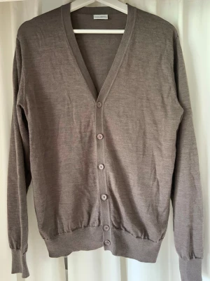 Merino cardigan John henric - Stilren beige kofta med v-ringning och långa ärmar. Koftan har fem knappar framtill och ribbade muddar vid ärmslut och nederkant. Tillverkad i ett tunt, mjukt material som passar perfekt för lager på lager.