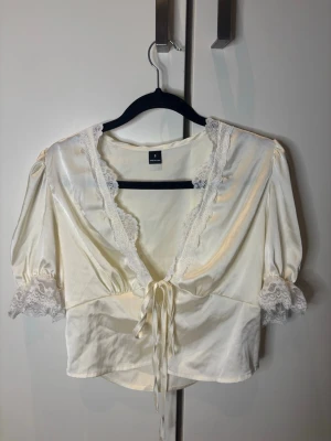 Vit kort blus med spetsdetaljer - Söt vit blus i glansigt material med korta puffärmar och spetsdetaljer längs urringningen och ärmsluten. Perfekt till både jeans och kjol. Oanvänd. 
