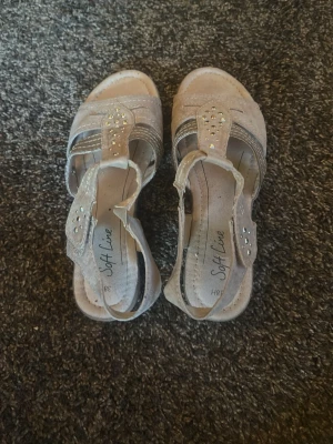 Beige sandaler med strass Soft Line - Säljer ett par beige sandaler från Soft Line med öppen tå och dekorativa strassdetaljer på remmarna. Sandalerna har en bekväm klack och elastiska band för enkel påtagning. Materialet ser ut att vara mockaimitation och de har en stilren design.