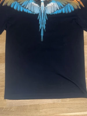 Svart t-shirt med färgglad fågelprint - Säljer en svart t-shirt från Marcelo Burlon med ett stort, färgglatt fågelmotiv i blått, orange och vitt över axlar och bröst. T-shirten har rund hals och korta ärmar. Perfekt för dig som vill sticka ut med en unik design.