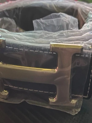Svart H bälte  - Snyggt svart bälte med ett stort H-format metallspänne. Bältet har vit kontrastsöm längs kanterna och är gjort i ett slätt material som liknar skinn. Perfekt accessoar för att lyfta din outfit.