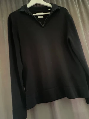 Svart merinoullströja med halvzip - Snygg svart stickad tröja från These Glory Days i mjuk superior merinoull. Tröjan har halvzip framtill, lång ärm och en clean, minimalistisk look som passar perfekt till jeans eller chinos.