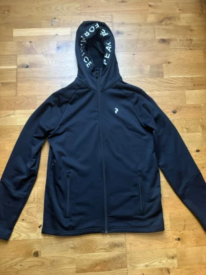 Svart hoodie med dragkedja och huva - Säljer en svart hoodie med dragkedja och två fickor framtill. Huvan har texten 'PERFORMANCE PEAK' tryckt på insidan och det finns en liten vit logga på bröstet. Materialet är mjukt och fleecefodrat på insidan för extra komfort. Storlek S
